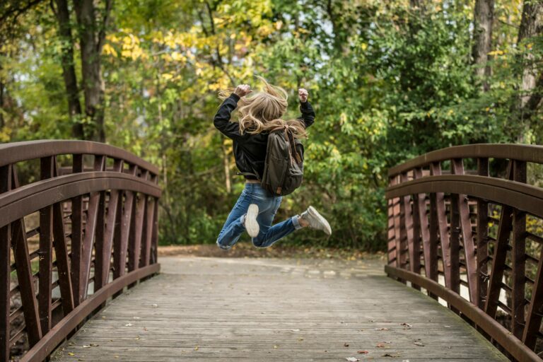 A blonde girl jumps exuberantly on a wooden bridge surrounded by autumn foliage, capturing the essence of joy and freedom.